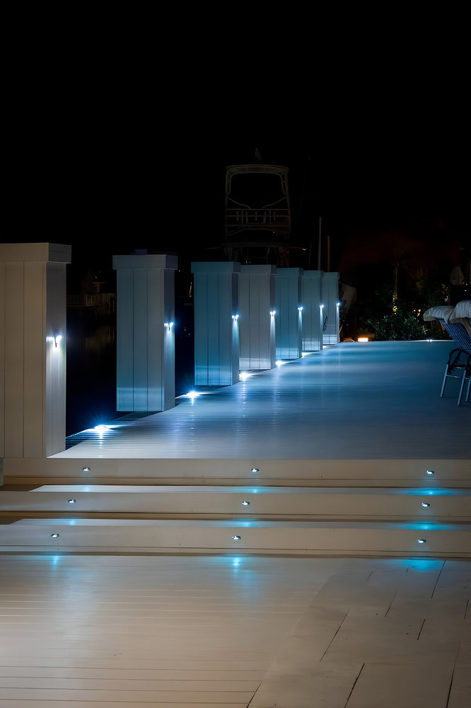 Luxury waterfront deck walkway at night with evenly spaced blue LED step lights and illuminated square piling caps — South Florida