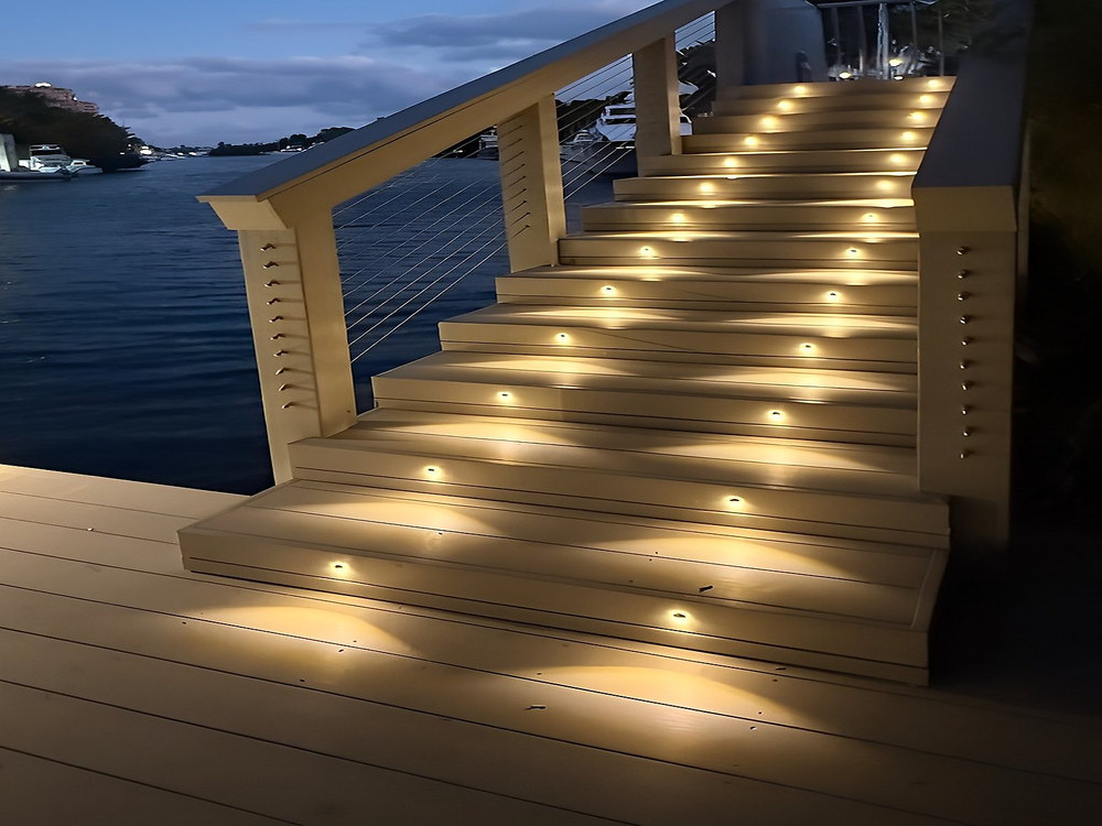 Waterfront composite deck staircase at twilight with warm golden LED step riser lights and cable railing — South Florida